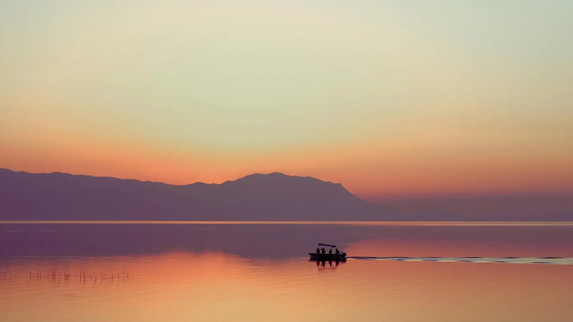Sunset over a calm lake with distant mountains and a fishing boat silhouette, evoking Utah Lake's flat waters and mountain backdrop